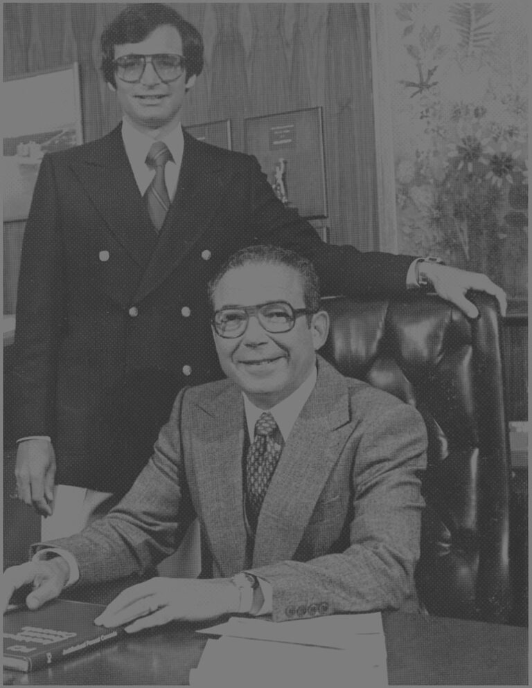 A vintage black and white photo of two men in an office setting. One man is seated at a wooden desk, wearing a suit with a tie and glasses, and smiling at the camera. The other man stands beside him, also in a suit with a tie and large glasses, resting one hand on the seated man's chair. In the background are framed pictures and a plant.