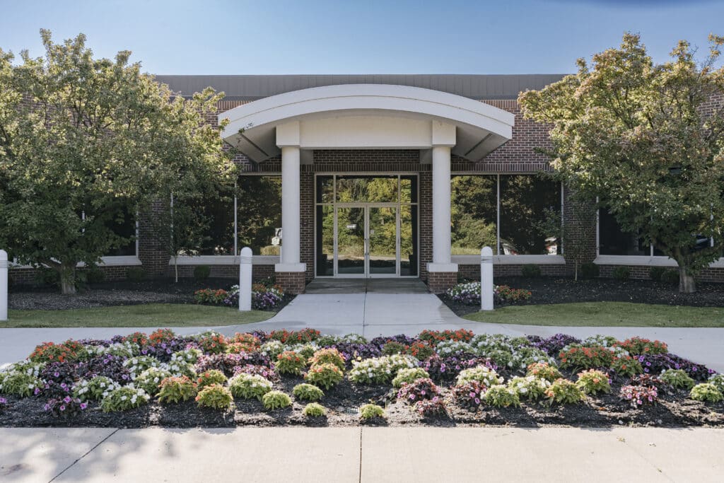 Entrance to an office building with a curved roof supported by pillars, surrounded by large windows. In the foreground, there is a well-maintained garden with colorful flowers and bushes, and two trees on either side of the entrance.