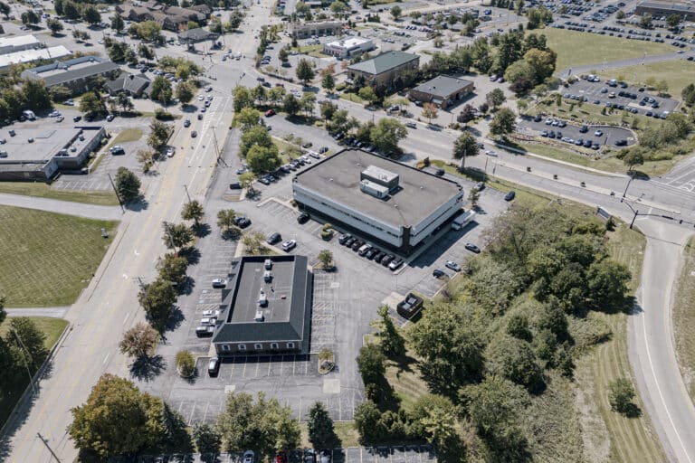 An aerial view of a suburban area featuring two large office buildings surrounded by parking lots. The area is bordered by roads with vehicles, and trees are scattered throughout the landscape. The scene captures a clear view of the organized urban layout, depicting both commercial and residential structures.