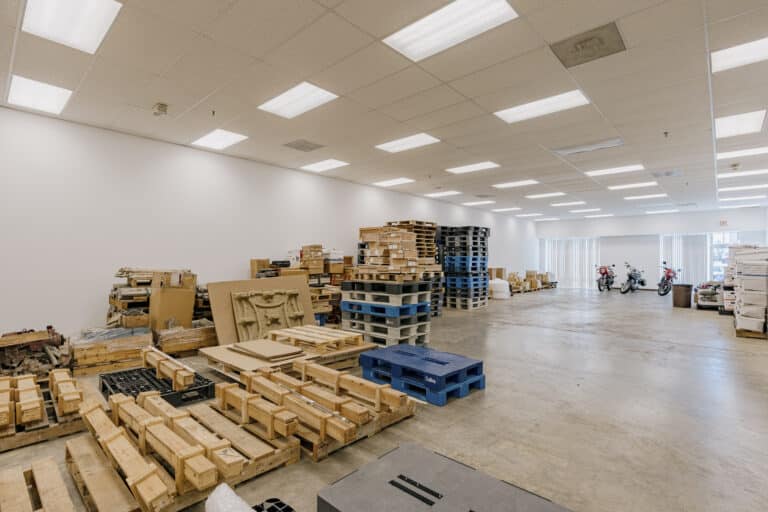 A spacious warehouse storage area with neatly stacked pallets, boxes, and packaging materials. The floor is concrete, and the ceiling is lined with fluorescent lights. In the background, motorcycles are parked near a set of large windows with vertical blinds, allowing natural light to filter in.