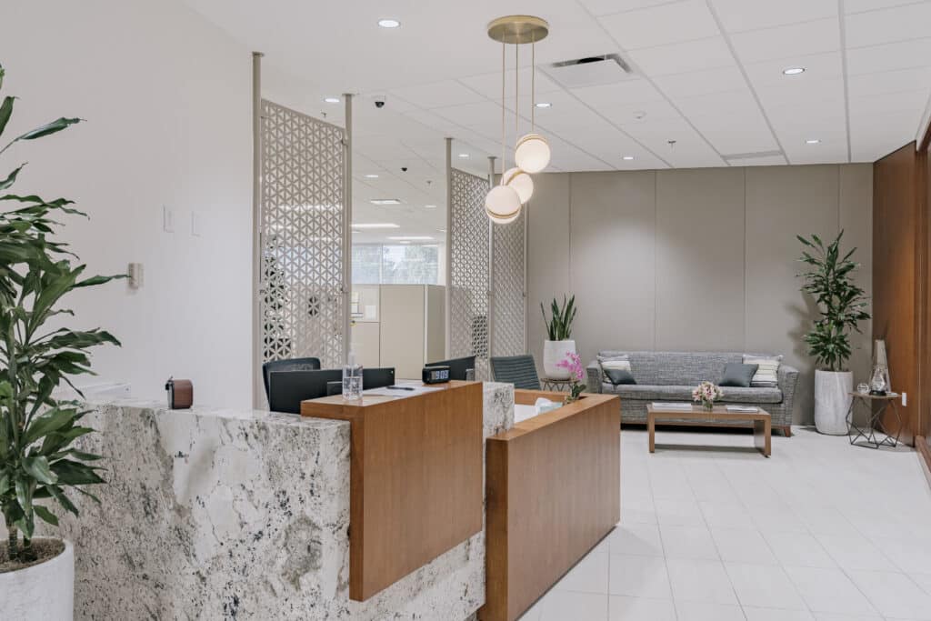 A modern office reception area featuring a marble and wood reception desk with a digital clock and hand sanitizer on top. The room includes a gray sofa with cushions, a wooden coffee table with a floral centerpiece, and decorative plants. The space has a light color scheme with white tile flooring, patterned room dividers, and stylish hanging lights.