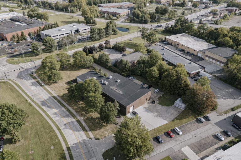 Aerial view of a suburban area featuring commercial buildings surrounded by greenery. A road with light traffic runs through the foreground, with parking lots and tree-lined paths visible around the structures. The landscape includes a mix of industrial and office buildings, with a backdrop of trees and open grass fields.