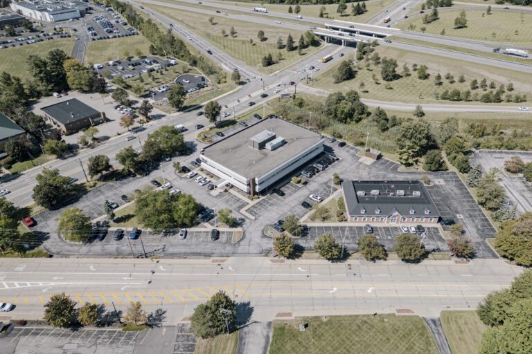 Aerial view of a commercial area featuring two office buildings with flat roofs surrounded by parking lots. The foreground shows a divided road with multiple lanes and marked crosswalks. In the background, an intersection connects to a highway overpass. Trees and green spaces are interspersed throughout the area.