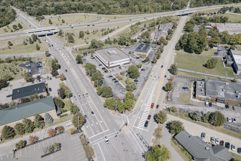 Aerial view of a modern urban intersection with multiple roads converging. Surrounding the intersection are several buildings with parking lots, and a highway overpass is visible in the background. There are numerous cars on the roads and parked in the lots. Green spaces and trees can be seen throughout the area, creating a mix of urban and natural elements.