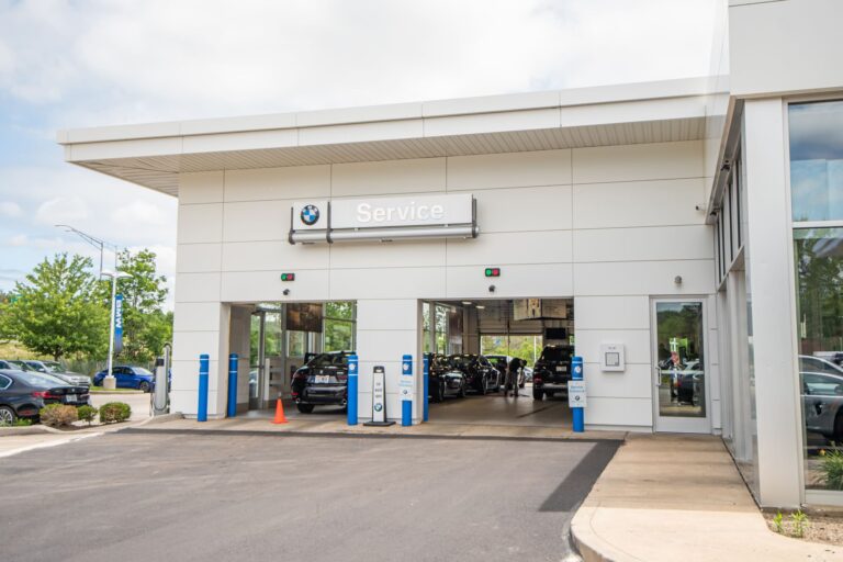 A BMW service center exterior with two open garage bays. Several cars, including BMW models, are parked outside. The building is modern with clean lines and features the BMW logo above a 'Service' sign. Trees and a clear sky are visible in the background.