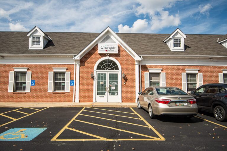 A single-story brick building with white trim and multiple windows, featuring a central arched doorway with a sign above it. Two cars are parked in a designated accessible parking area with clear markings in front of the building. The sky is partly cloudy.