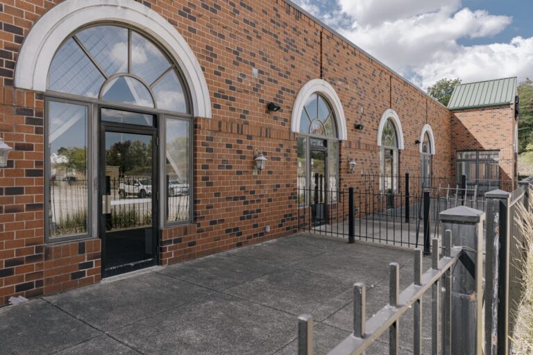 A brick building exterior featuring multiple large arched windows with a metal fence in front. The building has a modern design with red bricks and white accents, and there are outdoor lanterns mounted on the wall. The sky is clear with a few clouds, adding to the bright and open atmosphere.