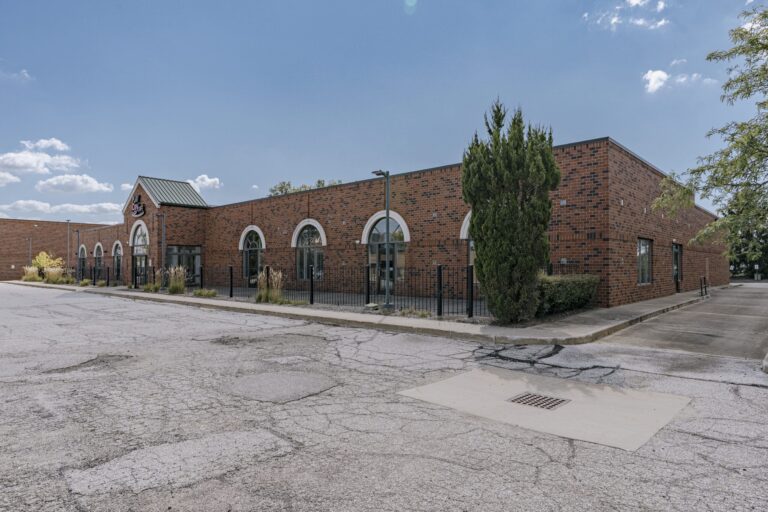 A large one-story brick building featuring multiple arched windows and a section with a green sloped roof. The building is surrounded by a black metal fence and various shrubs and trees. The foreground shows a wide, cracked asphalt surface under a clear blue sky with scattered clouds.