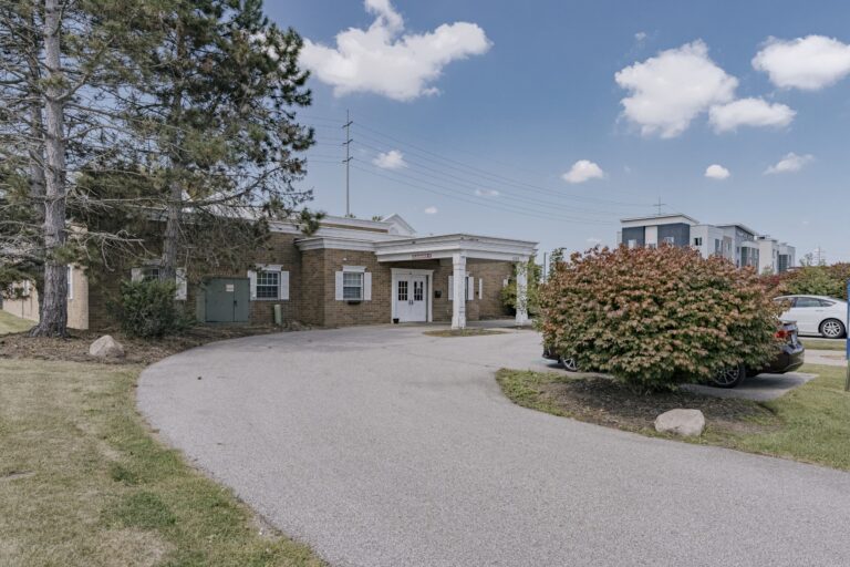A brick building with a prominent white entrance is surrounded by trees and a neatly trimmed bush. The sky is clear and blue with scattered white clouds. A curved driveway leads up to the building, and a few parked cars are visible in the background.