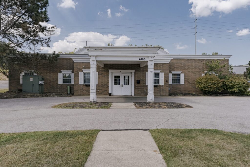A single-story brick building with white pillars at the entrance. The building has small windows with white shutters and a number 6100 displayed above the entrance. The foreground shows a concrete path leading to the entrance, surrounded by grass and a driveway. There are bushes on the right side and a tree on the left, with a few clouds visible in the blue sky above.