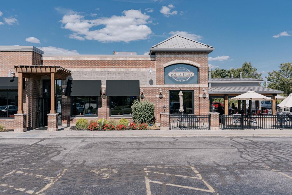 Front view of a brick building restaurant with a sign that reads 'Yours Truly Restaurant' above the entrance. The exterior features black awnings over large windows and a covered patio area with tables, chairs, and large umbrellas. The building is surrounded by a landscaped area with bushes and flowers under a clear blue sky.