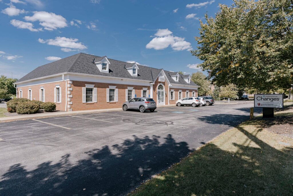 A single-story brick office building with a dark roof and white shutters is shown under a blue sky with scattered clouds. The building has several cars parked in front, with a paved parking lot and a grassy area with trees. A sign near the trees shows the name 'Changes' and an address.