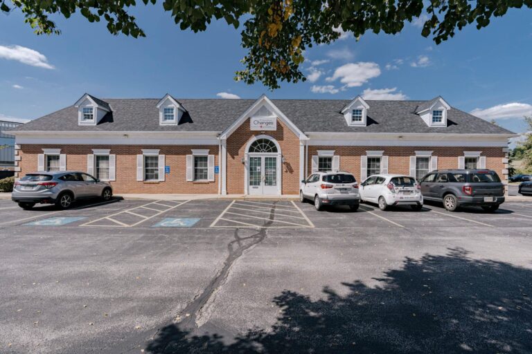 A single-story brick office building with large windows and a gabled roof. The entrance features a set of glass double doors and a sign above reading 'Changes'. There are several cars parked in the parking lot, with designated handicapped spaces visible in the foreground. The sky is clear with a few scattered clouds, and there are trees casting shadows on the parking lot.