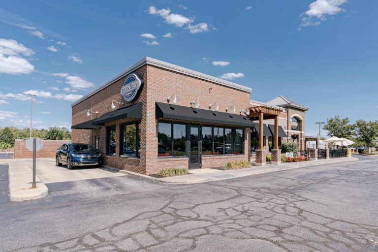 A brick restaurant building with large windows and a sign on the front, located in a parking lot under a bright blue sky. A black car is parked nearby, and the outdoor patio is adorned with plants and umbrellas.