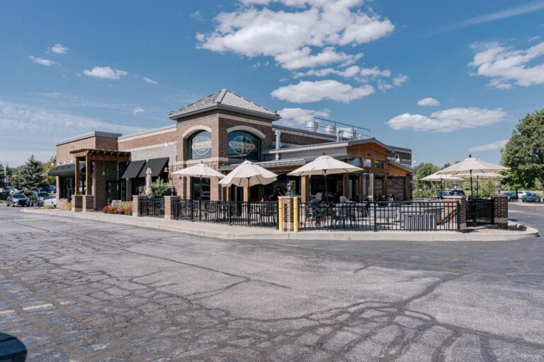 A brick restaurant with a small outdoor seating area featuring umbrellas over tables. The building has large windows and a sloped roof, with a parking lot in the foreground and a bright blue sky with scattered clouds above.