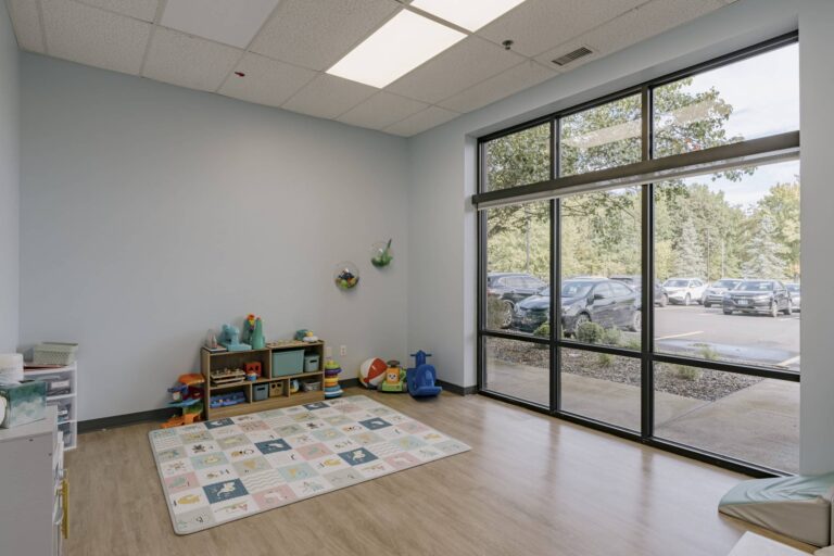 A bright playroom with a pastel-colored alphabet mat on a wooden floor. Shelves are filled with various colorful toys and bins against a light blue wall. Large windows on the right side allow natural light in, showing a view of parked cars and green trees outside.