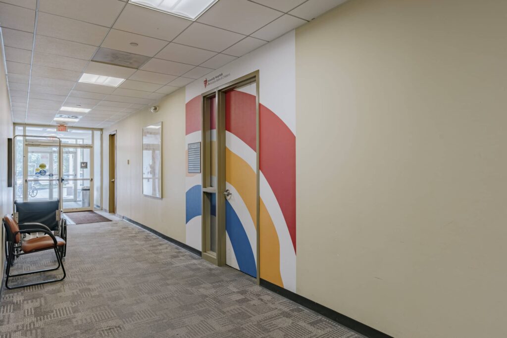 Interior view of a hospital hallway featuring a door with colorful painted arcs in red, yellow, and blue on the wall. The hallway has beige walls, grey patterned carpet, and a seated area on the left. Fluorescent lights are installed on the ceiling, and there are glass doors leading outside at the end of the hallway.