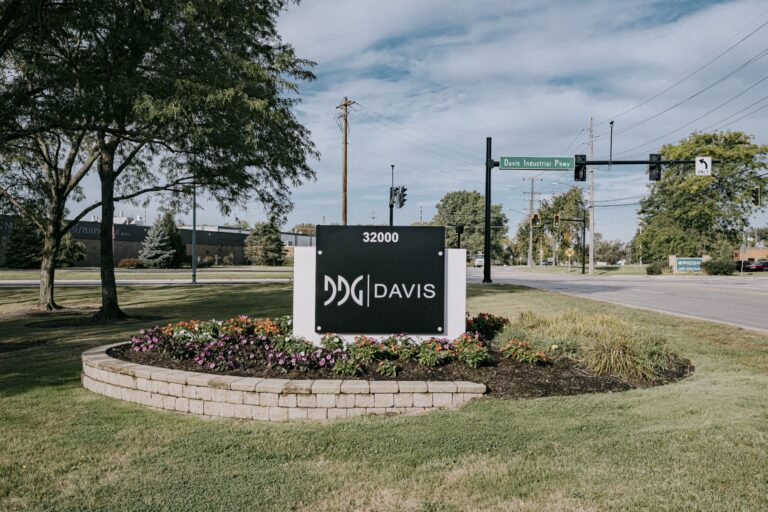 A Davis-branded sign with the number 32000 is prominently placed on a landscaped area with colorful flowers at the base. The sign is located near a street intersection with traffic lights and a green street sign labeled 'Davis Industrial Pkwy'. Trees and industrial buildings are visible in the background under a partly cloudy sky.