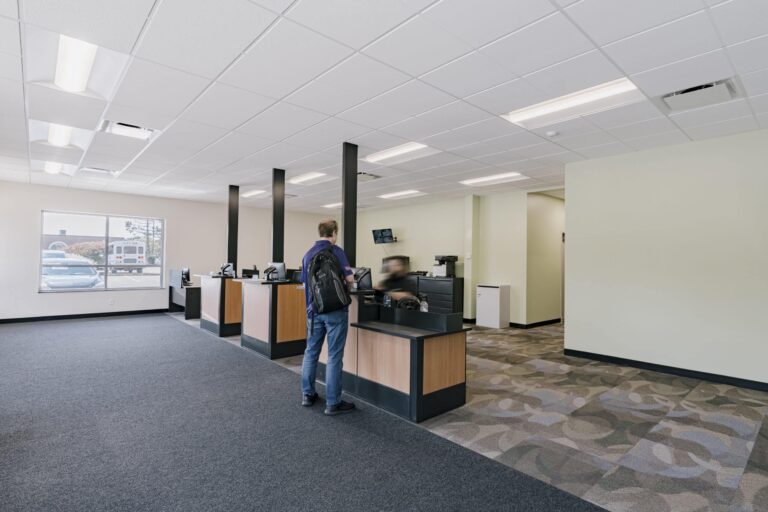 A modern office interior featuring multiple customer service counters with black and wooden finishes. A person wearing a backpack is standing at one of the counters, interacting with a staff member. The room has a patterned carpet, light-colored walls, and large ceiling lights. A window shows a view of parked vehicles outside.