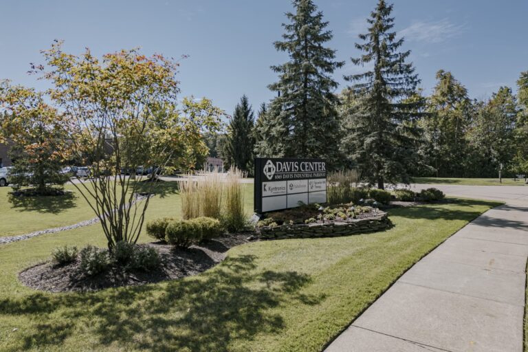 A well-maintained landscaped area featuring a sign for Davis Center located at 6656 Davis Industrial Parkway. The scene includes neatly trimmed grass, various bushes, and tall trees under a clear blue sky, with a sidewalk running along the right side.