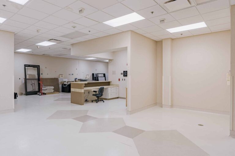 An empty hospital room undergoing renovation with a reception desk, some construction materials stacked, and a vacant office chair. The room has beige walls, a white and beige patterned floor, and a grid ceiling with fluorescent lights. A door frame and equipment trolley are visible against the wall.