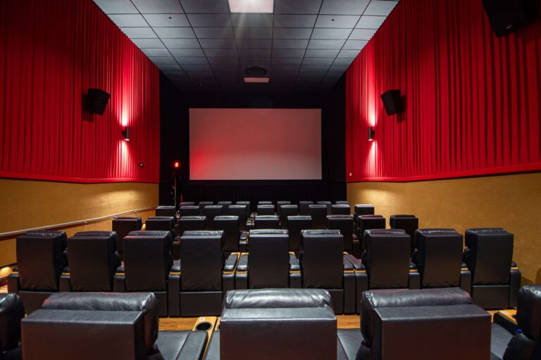 A modern cinema hall with red walls and a large white screen at the front. The seating consists of black leather recliners arranged in rows, providing a clear view of the screen. The walls are lined with red curtains and dim lighting fixtures, creating a cozy atmosphere.