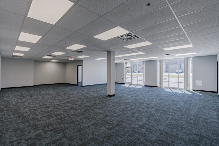 A spacious, empty office room featuring blue carpet, a grid of fluorescent ceiling lights, and large windows allowing natural light. The room has a grayish-blue wall with a black exit door, and the windows face a parking lot where a few cars can be seen.