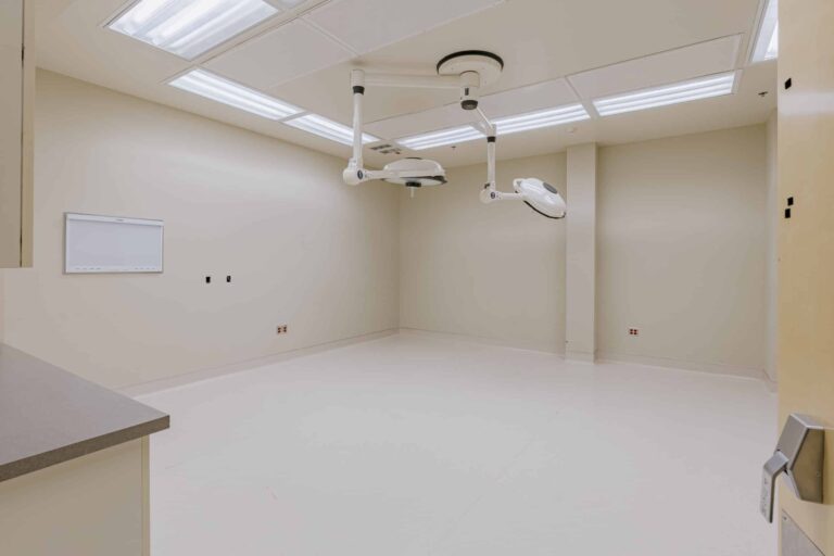An empty operating room with beige walls and white floors. Ceiling-mounted surgical lights are positioned at the center, and a blank whiteboard is visible on the left wall. Overhead fluorescent lights provide bright illumination.