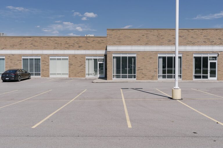 An empty parking lot in front of a single-story commercial building with a brick facade and large glass windows. A black car is parked to the left, and there are marked parking spaces and a tall light pole in the lot. The building appears to have no signage or people around, under a clear blue sky.