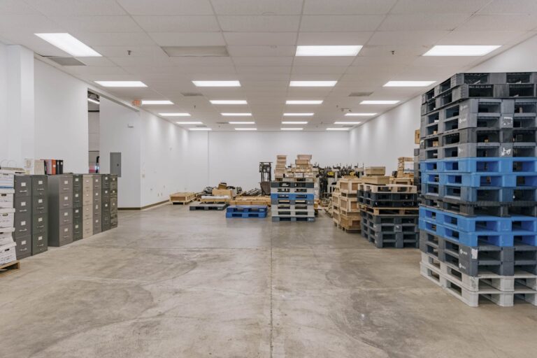 A spacious warehouse room with a polished concrete floor and illuminated by ceiling lights. To the left, there are rows of grey file cabinets with a few boxes on top. On the right, multiple stacks of blue and grey plastic pallets are neatly arranged. In the center, various wooden pallets and crates are scattered towards the back of the room.