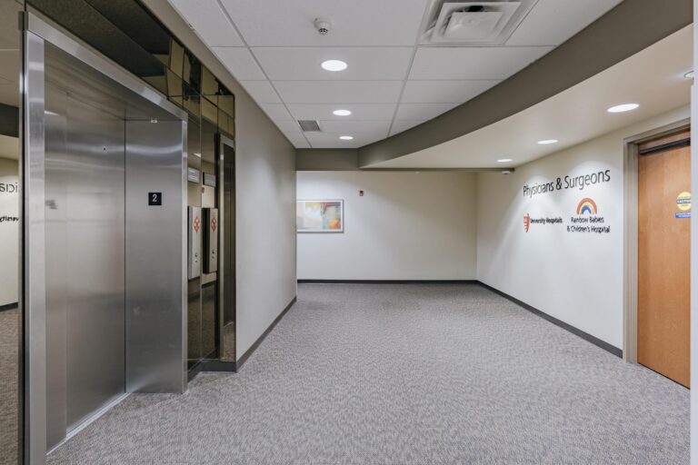 A hospital corridor featuring an elevator on the left with a number 2, and a hallway leading to a door labeled 'Physicians & Surgeons.' The wall displays logos for University Hospitals and Rainbow Babies & Children's Hospital. A piece of abstract art hangs at the end of the hallway.