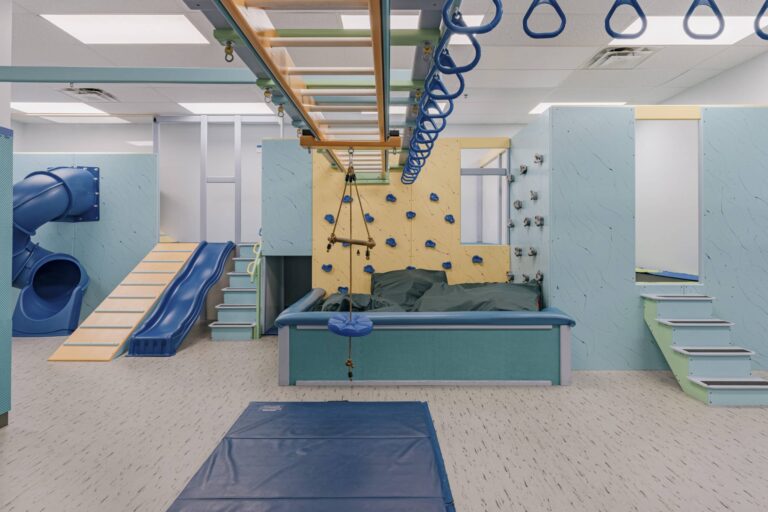 An indoor playground featuring a blue slide, a rope swing, and a climbing wall with blue holds. There is a padded area for safety, a climbing net, and a tube slide. The room has a light-colored floor and ceiling with fluorescent lighting.