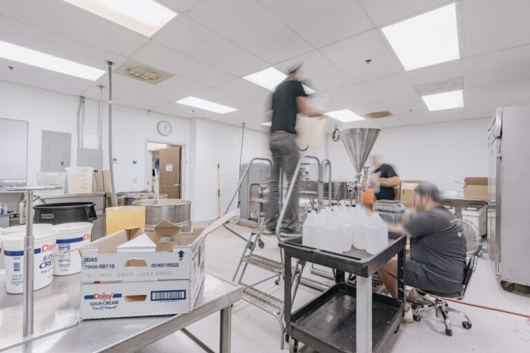 A busy industrial kitchen with workers engaged in tasks. One person stands on a step ladder pouring contents into a large funnel, while another person manages bottles on a rolling cart. The room is equipped with stainless steel tables and containers. Boxes labeled 'Daisy Sour Cream' are visible in the foreground. The kitchen is well-lit with large ceiling lights and a clock on the wall shows the time.