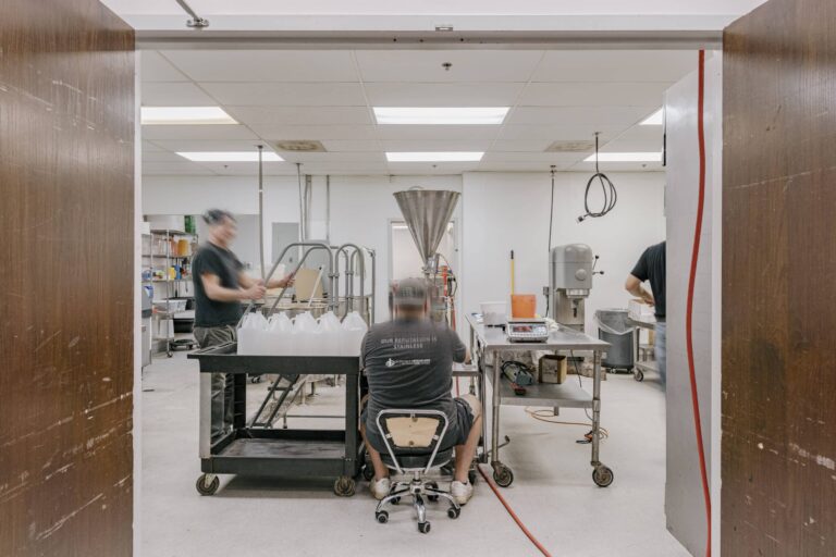 Image of a commercial kitchen where multiple workers are engaged in tasks. One person is seated on a chair next to a stainless steel table with various equipment, while another person stands beside a cart filled with plastic containers. The workspace includes machinery, hanging cables, and a bright overhead lighting.