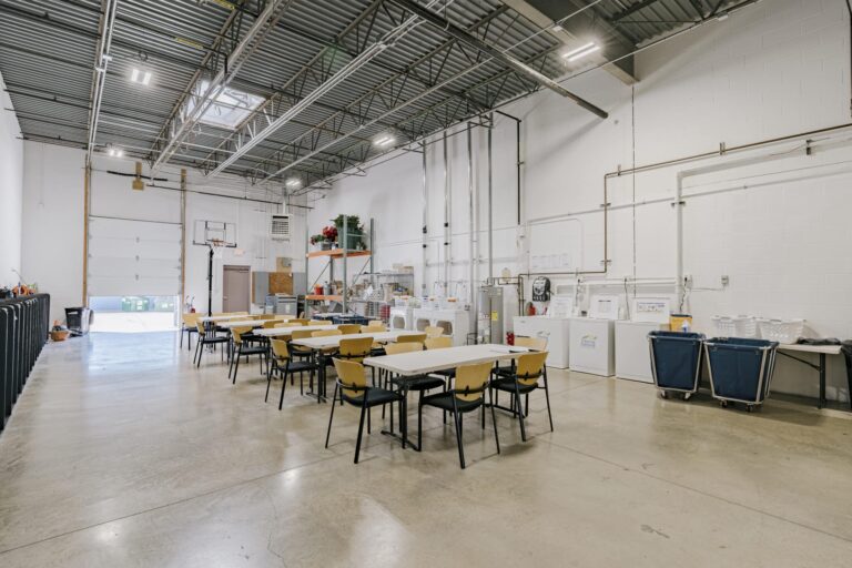 A spacious industrial room with a high ceiling and visible metal ductwork. The area includes multiple tables and chairs, shelving with various items, washing machines, large blue laundry carts, and a basketball hoop near a large garage door, allowing natural light into the space.