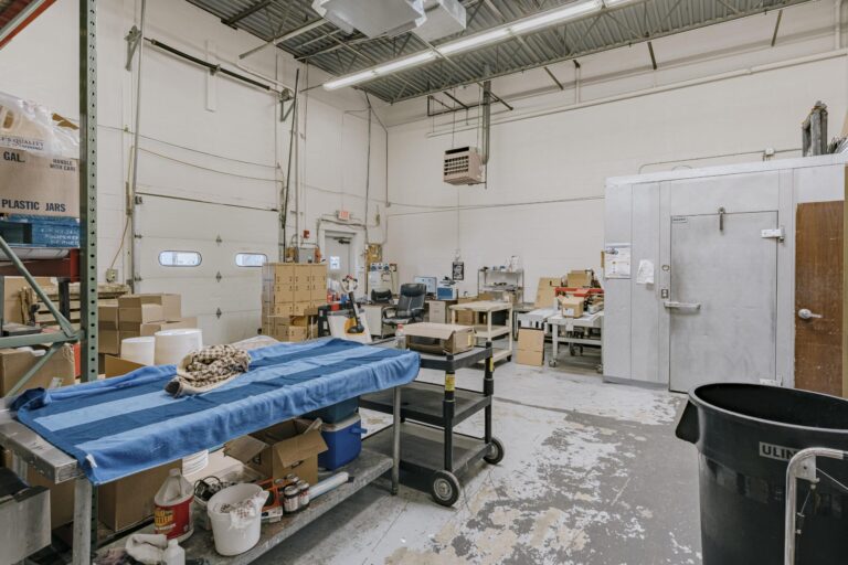 Interior of an industrial storage room featuring a blue-covered table surrounded by various boxes and supplies, including a large trash bin, shelving, and a metallic storage unit. The space has high ceilings, fluorescent lighting, and various equipment and cardboard boxes scattered throughout the room.