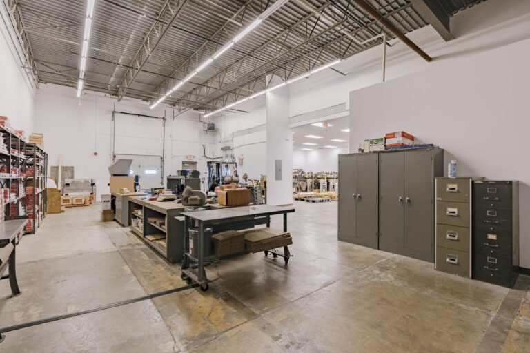 Spacious industrial warehouse interior with high ceilings, storage shelves filled with boxes, work tables with various items, and metal file cabinets against a wall. A bright adjoining room with sewing machines on pallets is visible in the background.