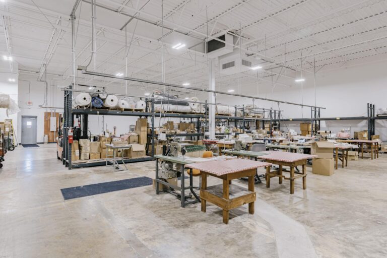An industrial workspace with large tables covered in patterned fabric, surrounded by storage racks filled with rolls of materials and boxes. The space is well-lit with bright ceiling lights and has a clean, organized appearance with equipment and materials neatly arranged.