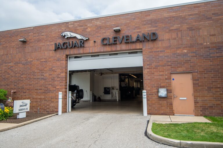 Brick building with signage reading 'Jaguar Cleveland' above a large garage door that is partially open, revealing a vehicle service area inside. A small sign to the left of the entrance displays 'Jaguar Service'. The scene includes a side door and some landscaping with flowers and grass.