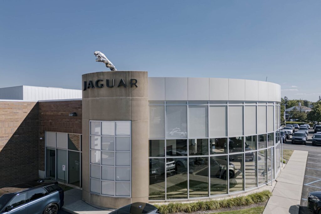 A Jaguar dealership featuring a cylindrical building with large glass windows and a prominent Jaguar logo on a beige column. A silver jaguar statue is mounted on top of the column, and parked cars are visible around the building under a clear blue sky.