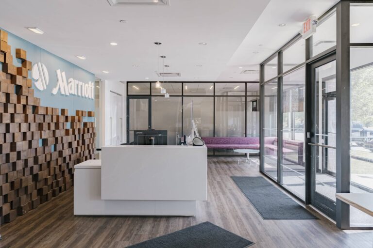 A modern hotel reception area with a white desk and a decorative wooden wall displaying the Marriott logo. The room features wooden flooring, large glass windows allowing natural light, and a waiting area with pink seating. An exit sign is visible above the glass doors.