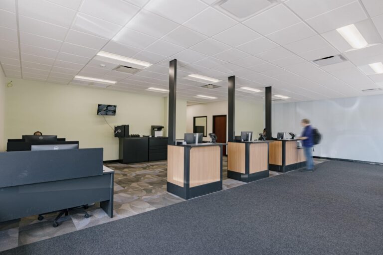 A modern bank interior with four wooden and black teller counters lined up, each equipped with computers and other equipment. The space has a light green wall and a carpeted floor. A person wearing a backpack is seen walking through, and there are monitors and office equipment in the background.