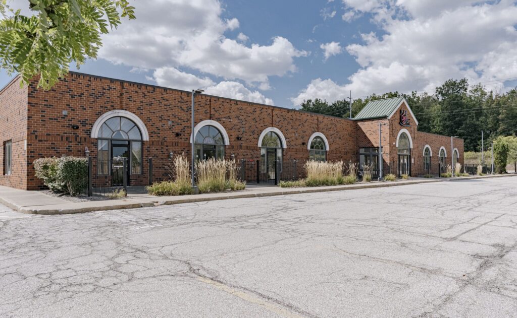 A modern brick building featuring arched windows with white trim, surrounded by landscaped greenery and tall ornamental grasses. The building has a green roof section, and the sky above is blue with scattered clouds. The parking lot in front is empty and shows some wear.