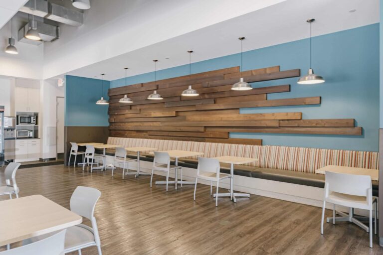 A modern cafe interior featuring a long wood-paneled wall with several overhead pendant lights. There is a row of cushioned seating with striped upholstery along the wall, accompanied by light-colored tables and chairs against a wooden floor.
