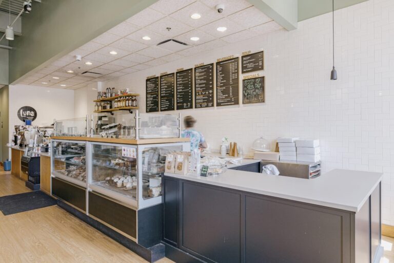 The image shows the interior of a modern cafe with a light wood floor and white tile walls. There is a glass display case on the counter featuring baked goods like cupcakes and pastries. Behind the counter, a person in a colored shirt is busy, and a menu board on the wall lists various food and drink options. Shelves above the counter hold bottles and jars, while a sign on the wall displays a special offer for vegan chili.