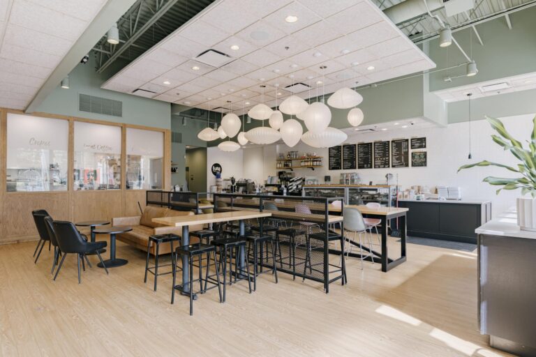 A modern cafe interior featuring tables with chairs and stools, light wooden flooring, and a ceiling adorned with artistic white paper lanterns. The seating area has a mix of black chairs and a brown leather bench. The bar area is visible with menu boards overhead, a counter with a cash register, and shelves stocked with coffee supplies. Natural light enters from large windows on the left, and a tall green plant is seen in the foreground on the right.