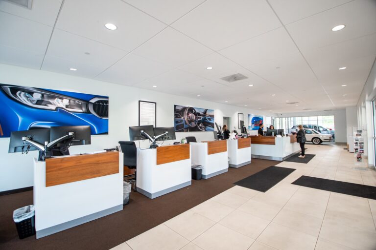 Interior of a modern car dealership featuring several service desks with computer monitors and chairs. Large car images are displayed on the white walls above the desks. Two people are seen interacting with staff members at the service counters, and a car is visible through the large glass windows in the background.