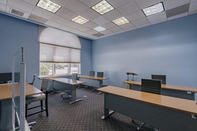 A modern classroom with light blue walls and several wooden desks arranged in rows. Black swivel chairs are placed at each desk, and overhead lighting illuminates the room. A large window with partially drawn blinds allows natural light to enter. The room is empty, suggesting it is ready for a class or meeting.