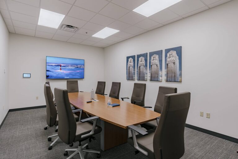 A modern conference room featuring a long wooden table surrounded by eight brown high-back office chairs. A large digital display screen is mounted on the wall showing a scenic image with text prompting for Zoom control. Four artistic panels of statues are hung on the opposite wall. The room has a carpeted floor and a white ceiling with recessed lighting.