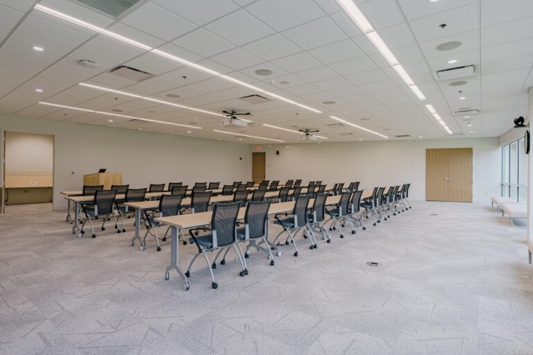 A spacious, modern conference room with multiple rows of tables and mesh office chairs, fluorescent ceiling lights, a projector, and a podium at the front. The room has a light-colored carpet and large windows on one side, with wooden doors at the back.